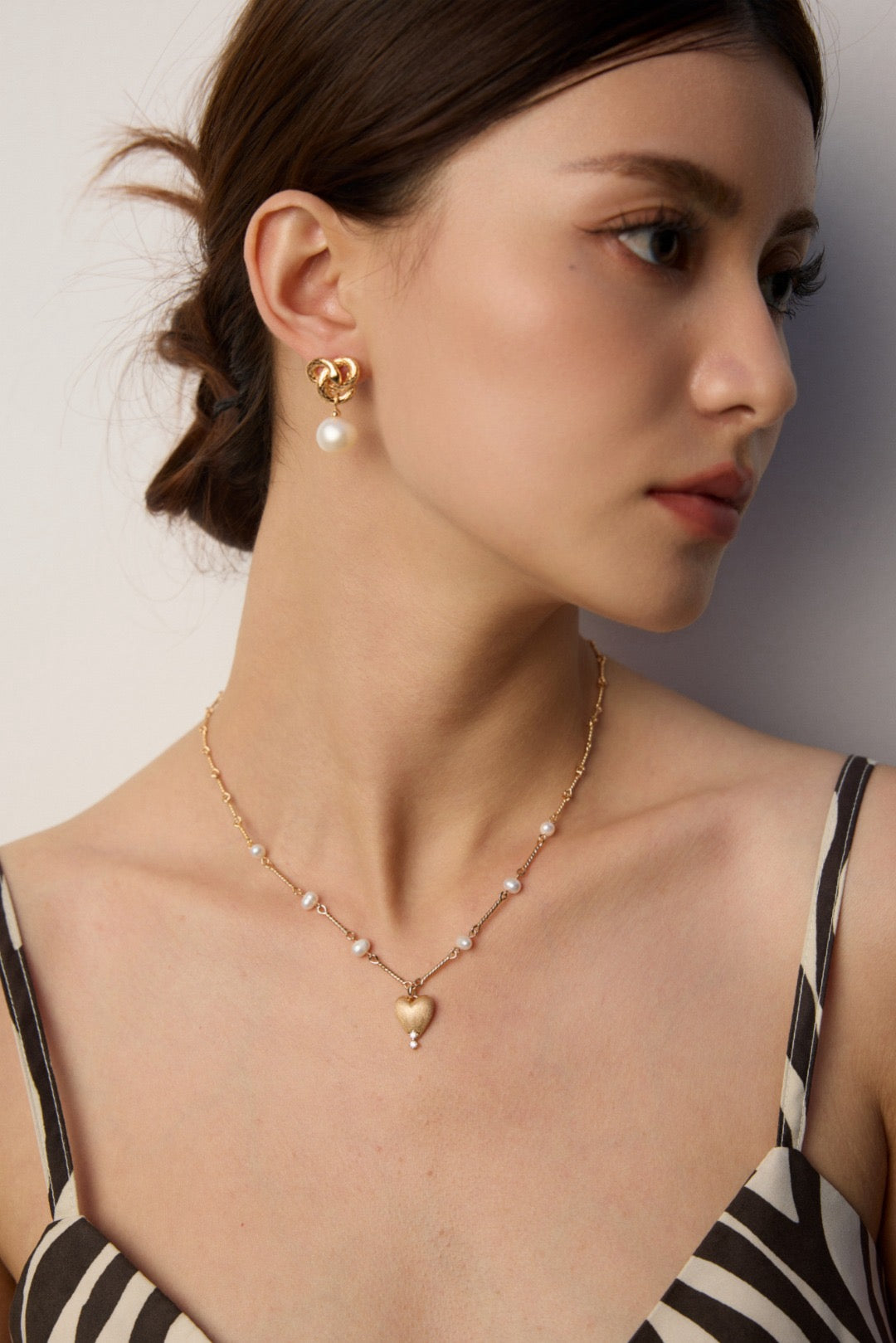 Woman wearing a gold necklace with heart pendant and pearl earrings against a neutral background