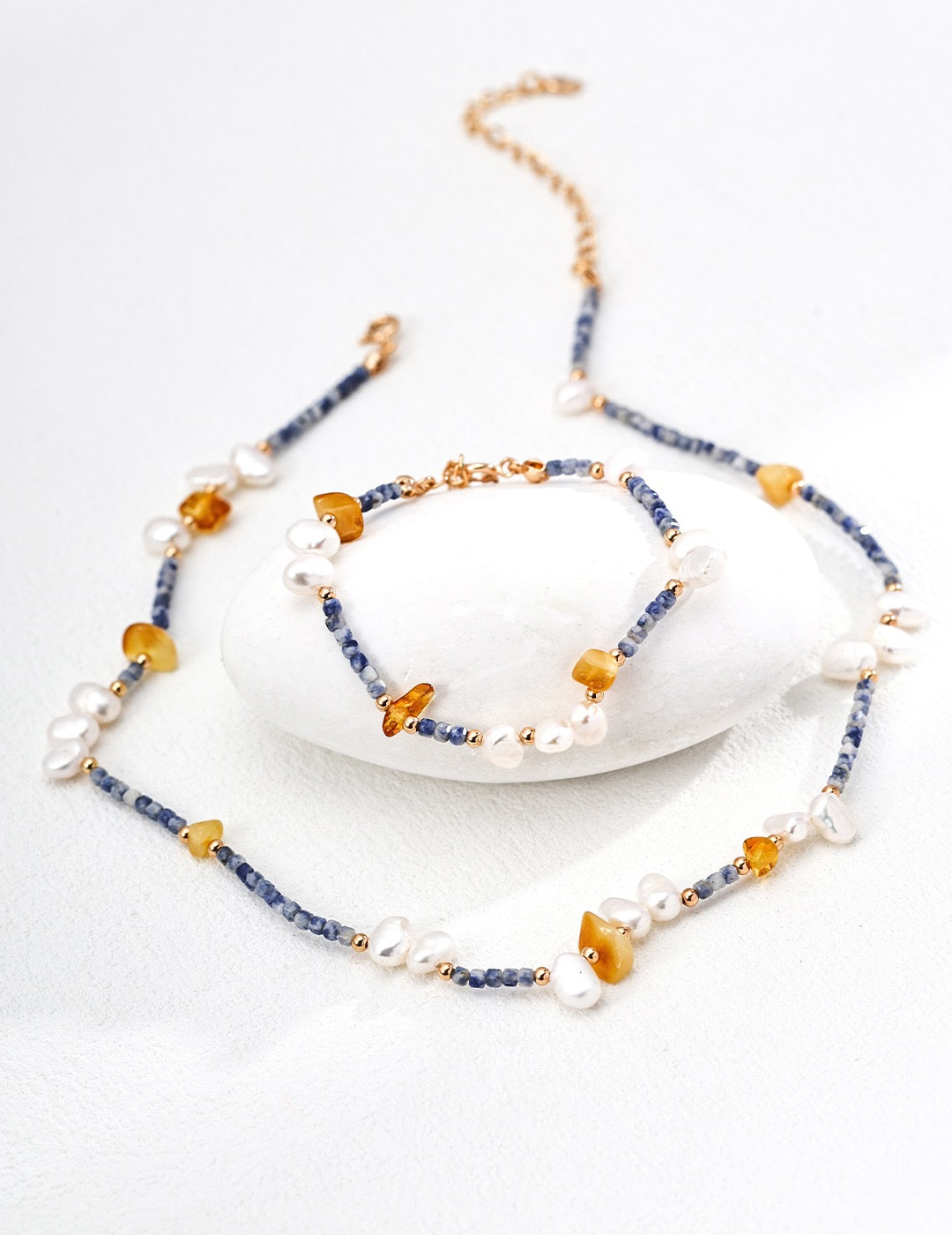 A delicate jewelry set featuring a necklace and bracelet made of irregular freshwater pearls, honey-toned amber stones, and deep blue sodalite beads, accented with tiny gold spacers. The pieces are displayed elegantly on a smooth white stone and light background.