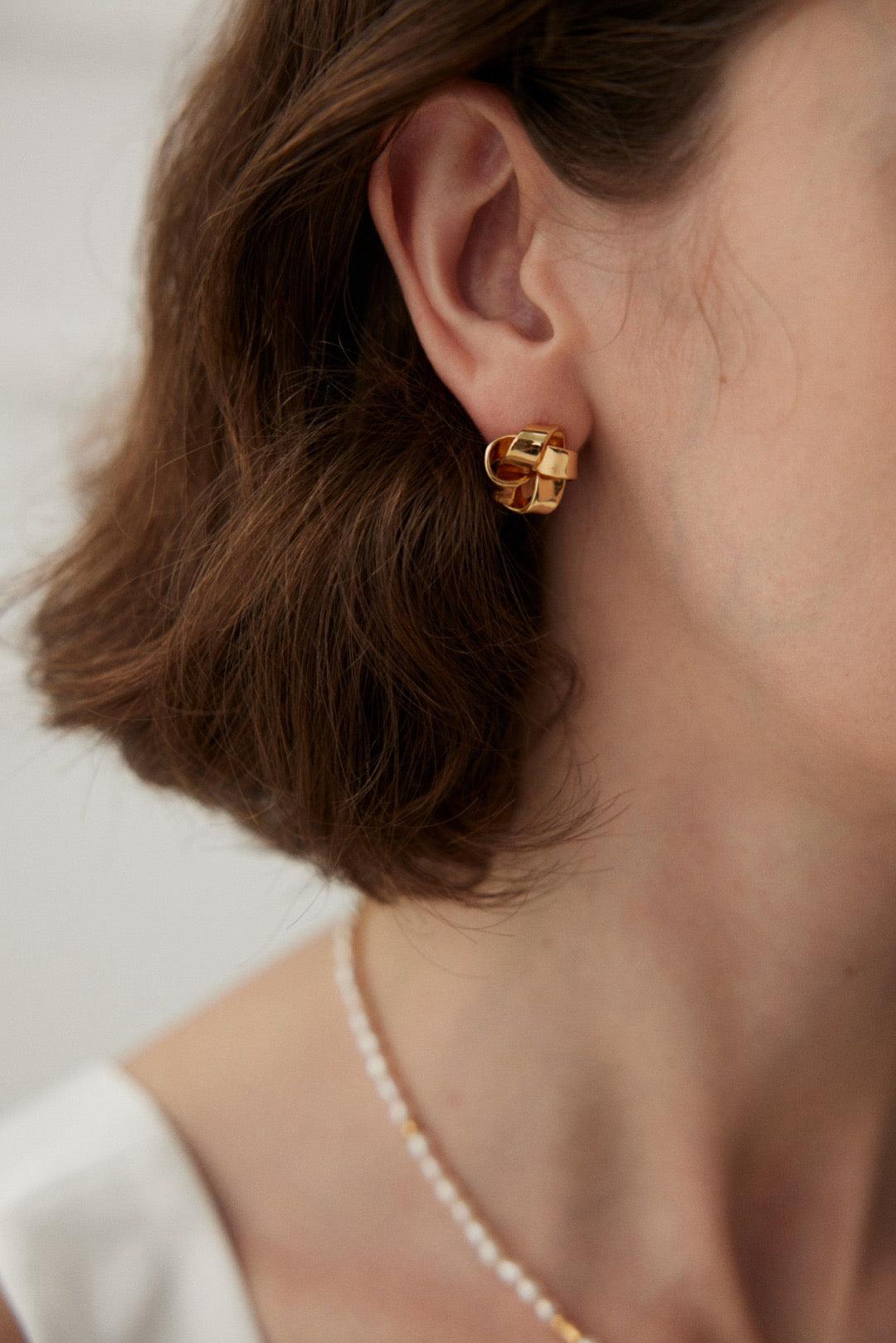A person wearing the Anissa Earrings, which are gold stud earrings with a knot design.