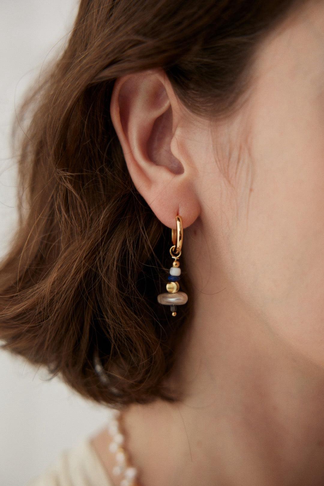 A close-up image of a woman's ear wearing an asymmetric earrings with gold, pearl, and blue gemstone details.