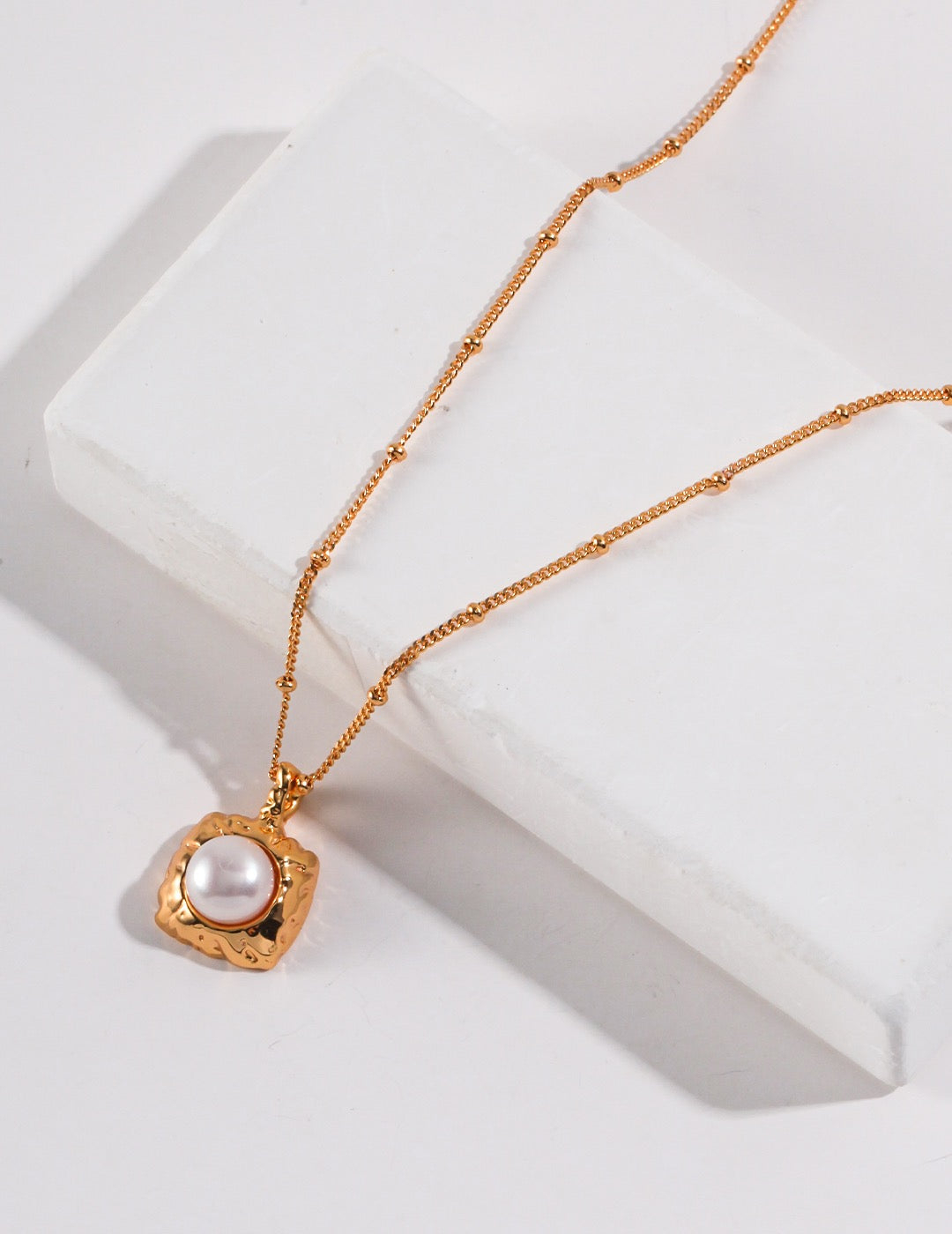 Gold necklace with a square-shaped pendant featuring a pearl on a white surface