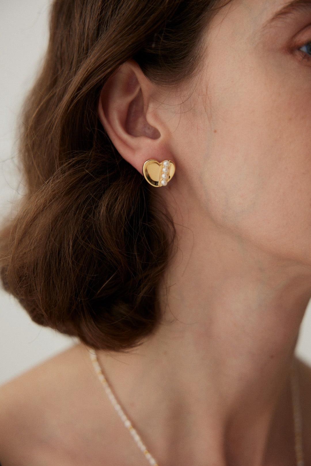 A close-up image of a woman's ear wearing a gold vermeil stud earring with a heart shape and shell design.
