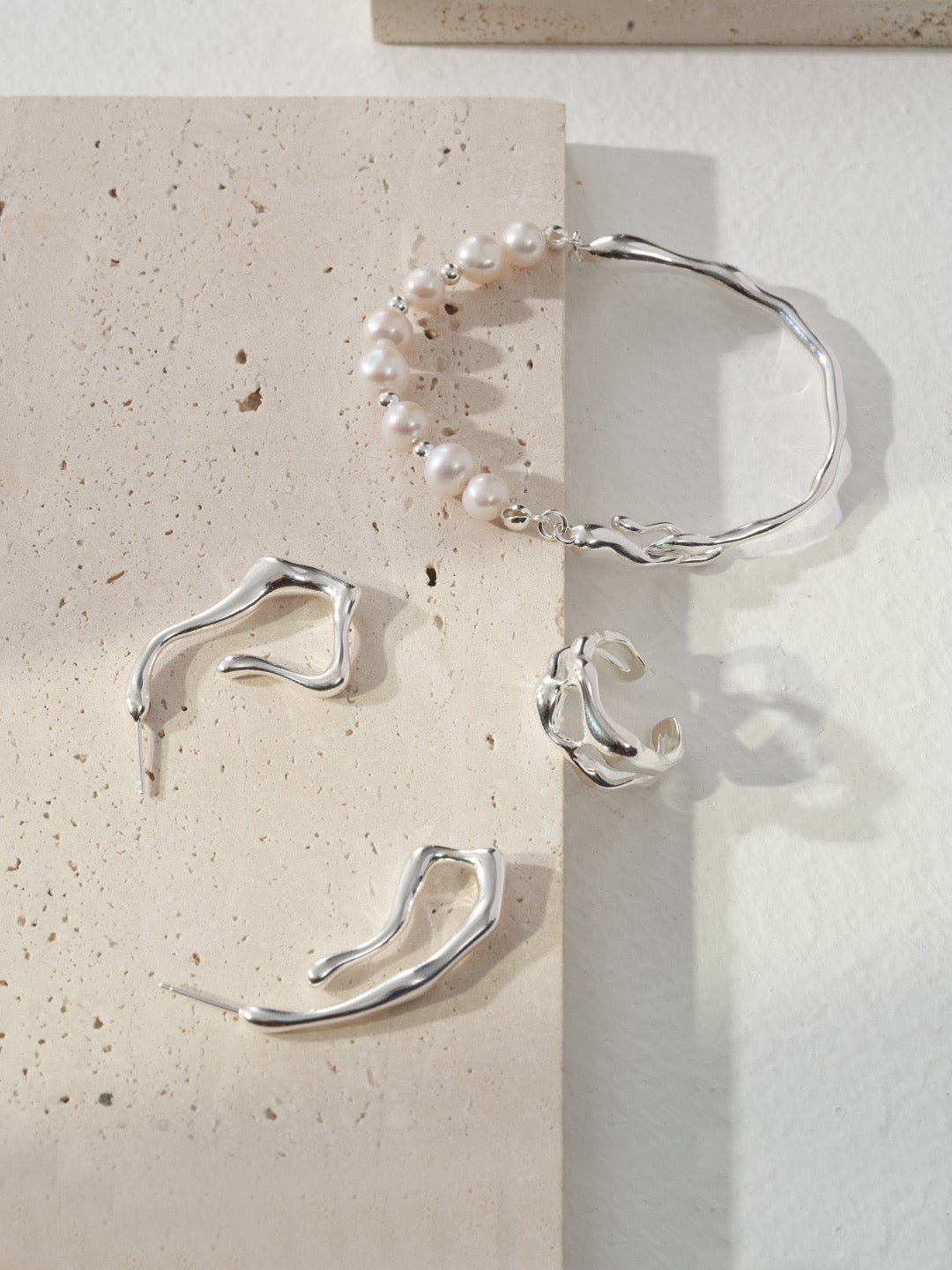 Caitlin Minimalist Pearl Asymmetrical Bracelet In 18K Gold and silver and three pieces of silver jewelry are displayed on a marble surface with soft lighting.