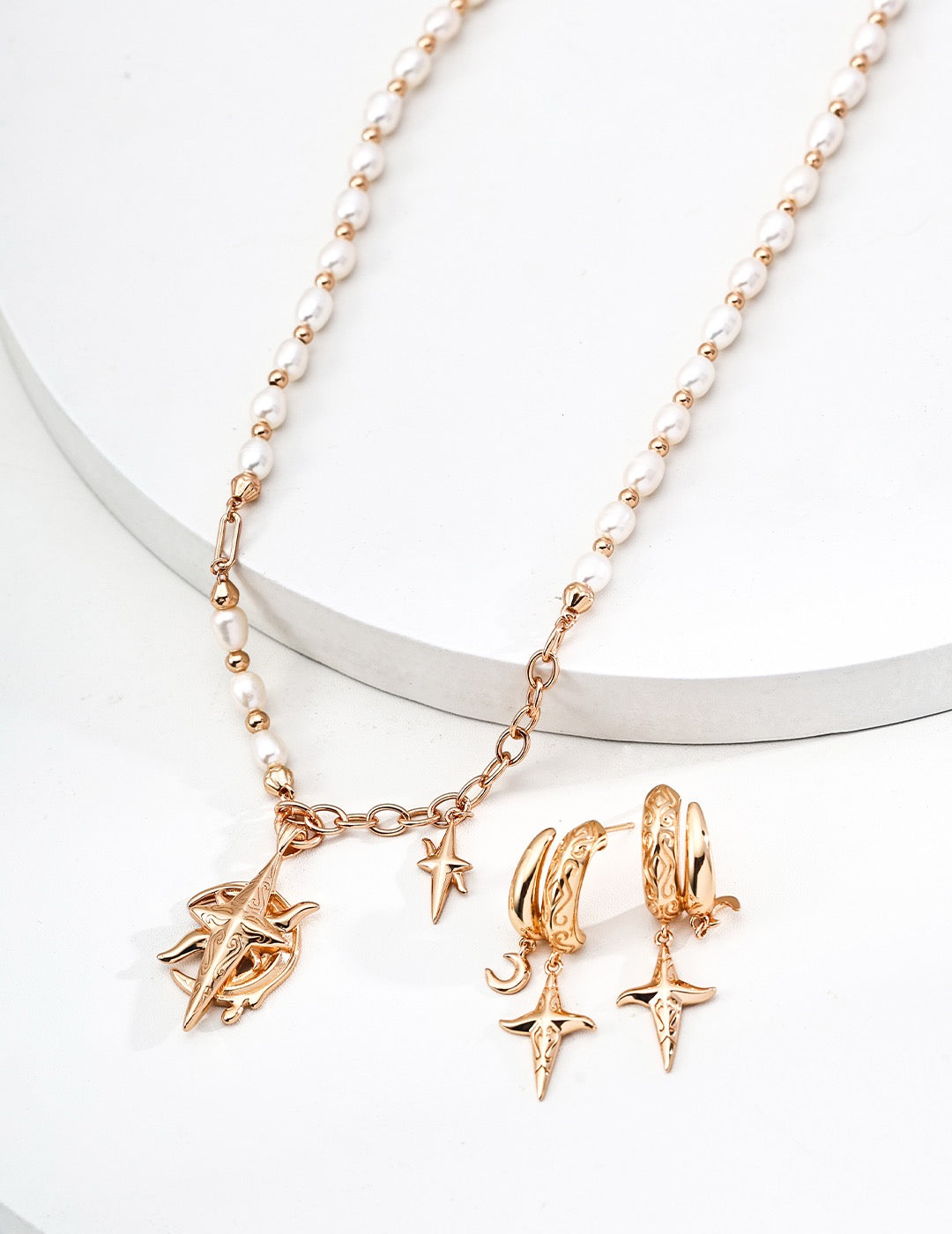 Gold and pearl jewelry set featuring a charm necklace with celestial motifs and matching engraved hoop earrings, styled on a white surface.