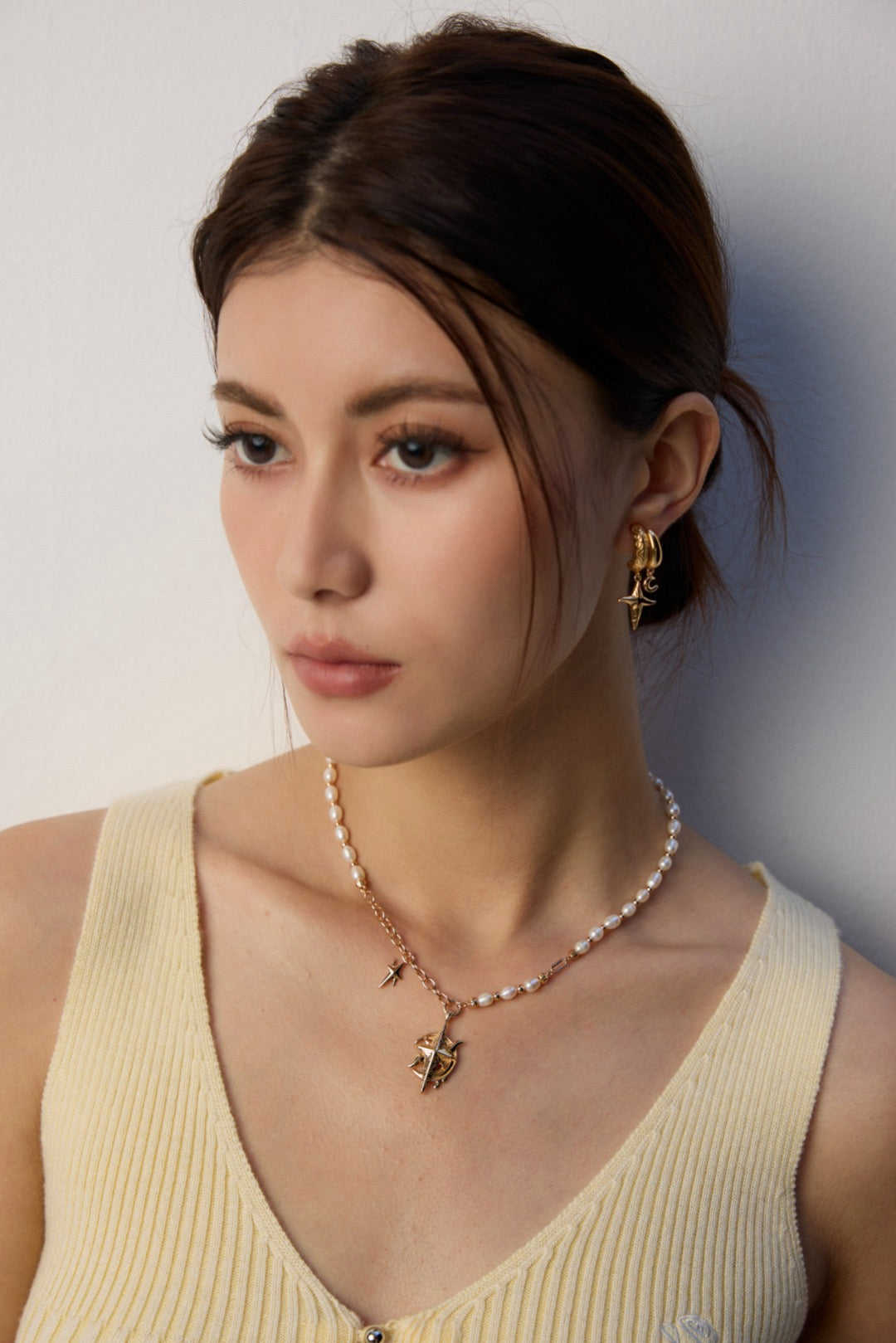 The model is wearing the Celestial Crest Pearl Necklace and the hoop earrings in a yellow knit tank top.