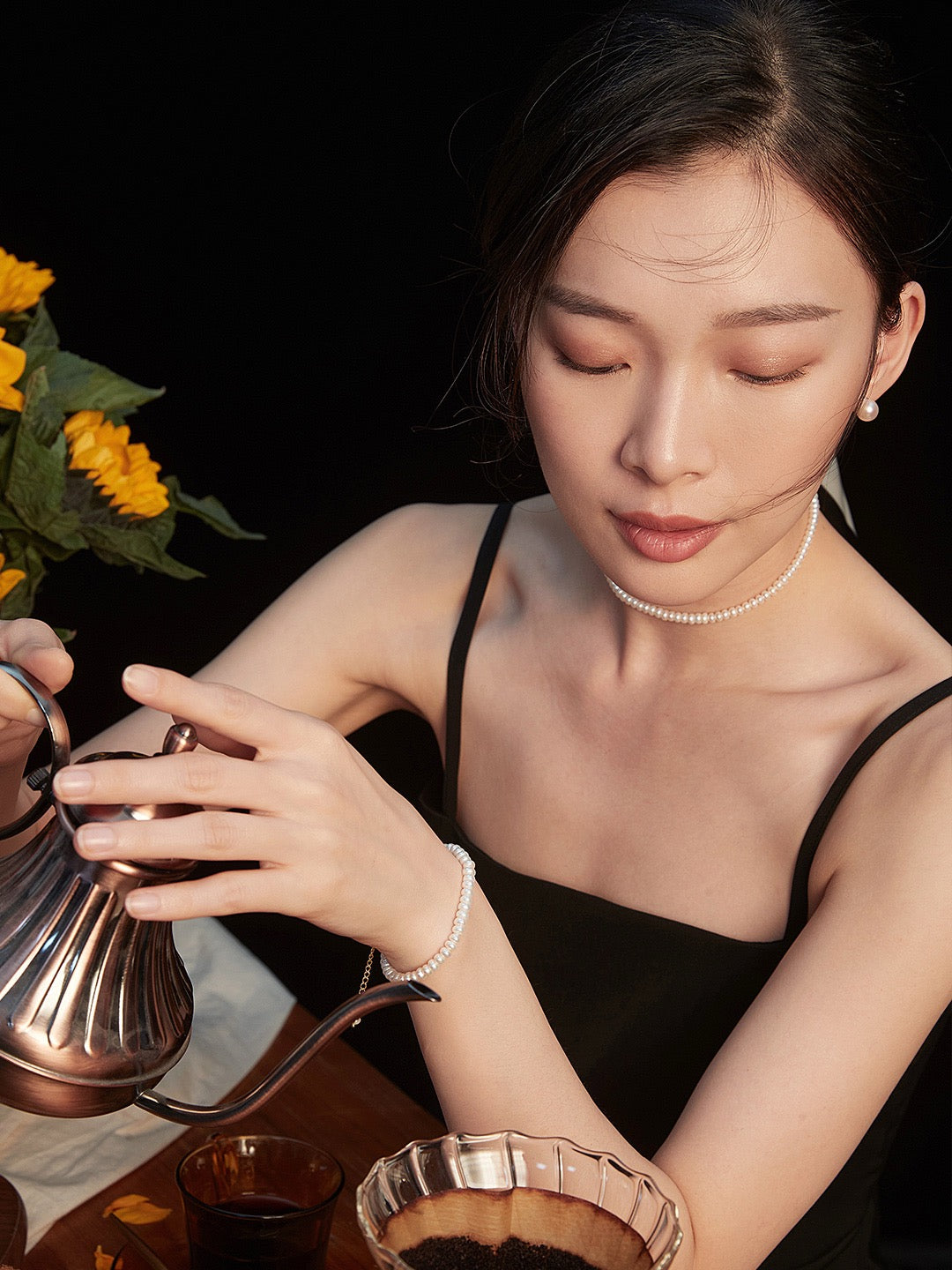 Woman is wearing pearl stud earrings and pearl choker necklace and she is pouring tea from a silver teapot with sunflowers in the background