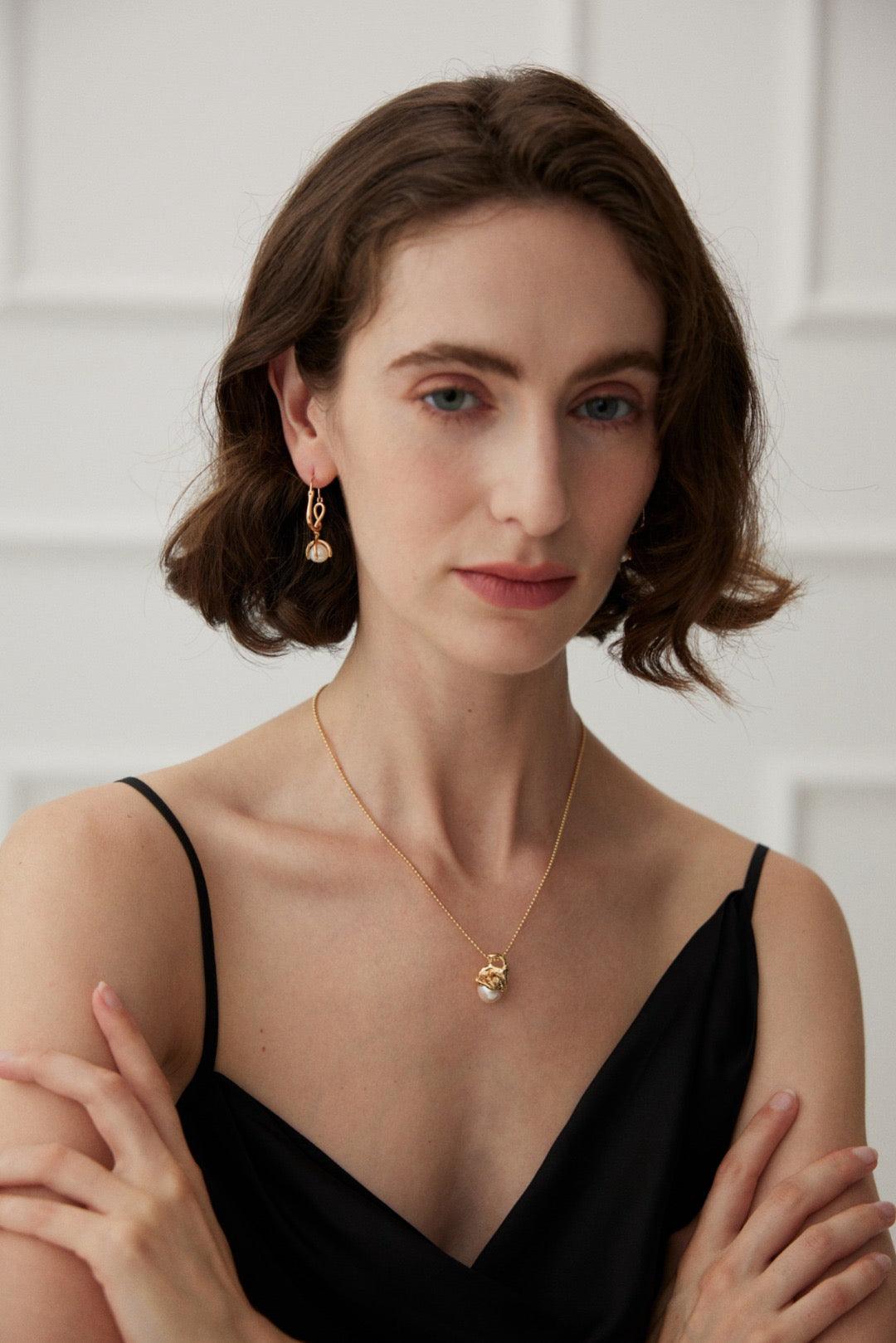 A person wearing a black V-neck top showcasing a gold pendant necklace with a pearl in the center against the neutral background.