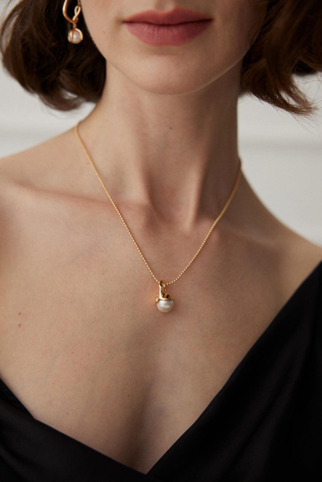 A person wearing a black v-neck top showcasing a gold pendant necklace with a pearl in the center.