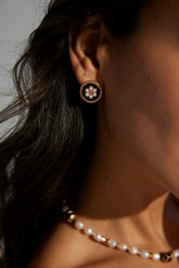 A close-up image of a woman's ear wearing a gold-colored earring with a black and white pattern, accompanied by a gold chain necklace.
