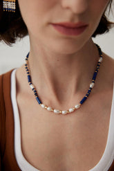 A person wearing a white top and brown accessory, showcasing a necklace with blue (Lapis Lazuli) and white (pearl) beads.