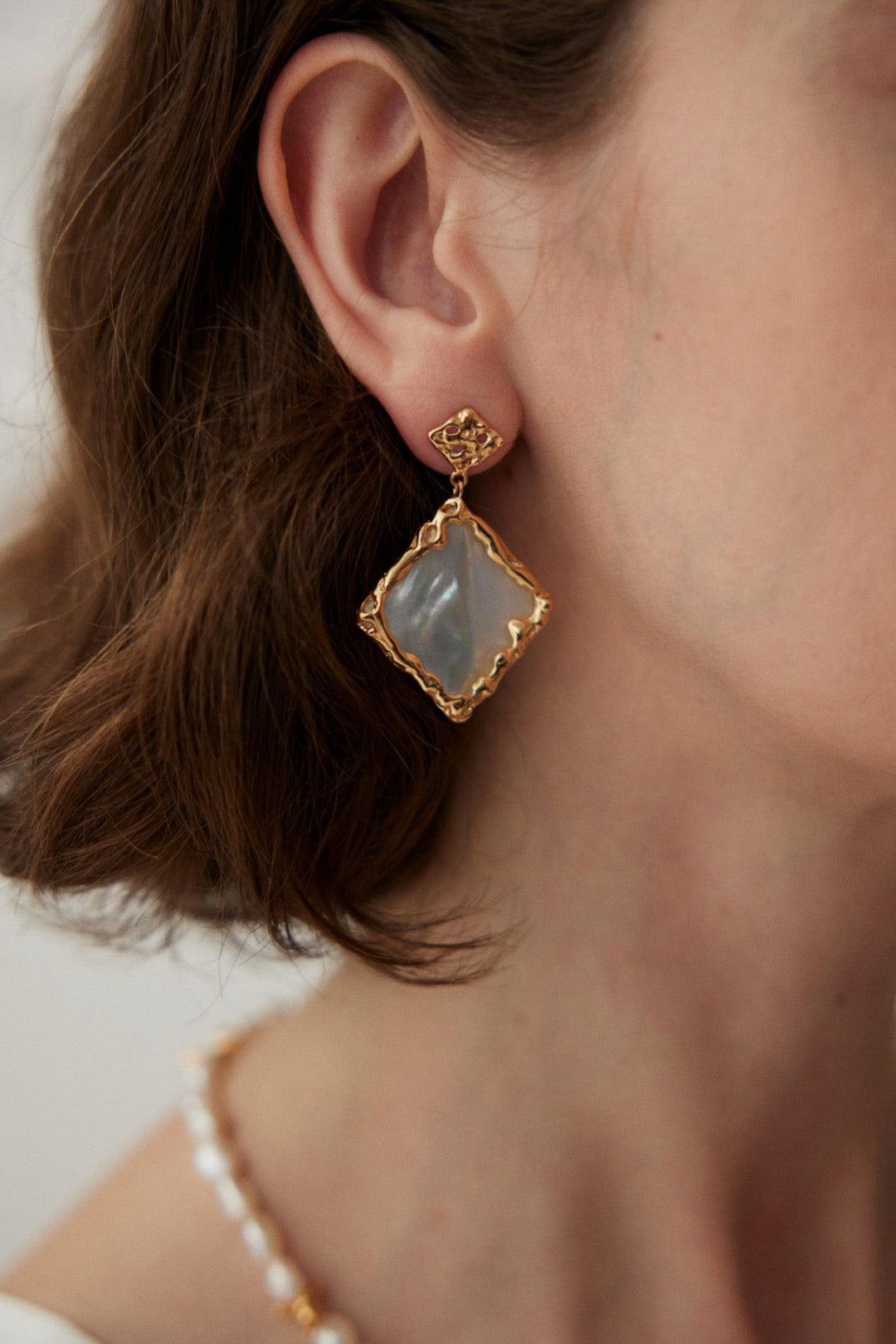 A close-up image of a woman's ear wearing a gold vermeil earring with a square-shaped mother-of-pearl inset.