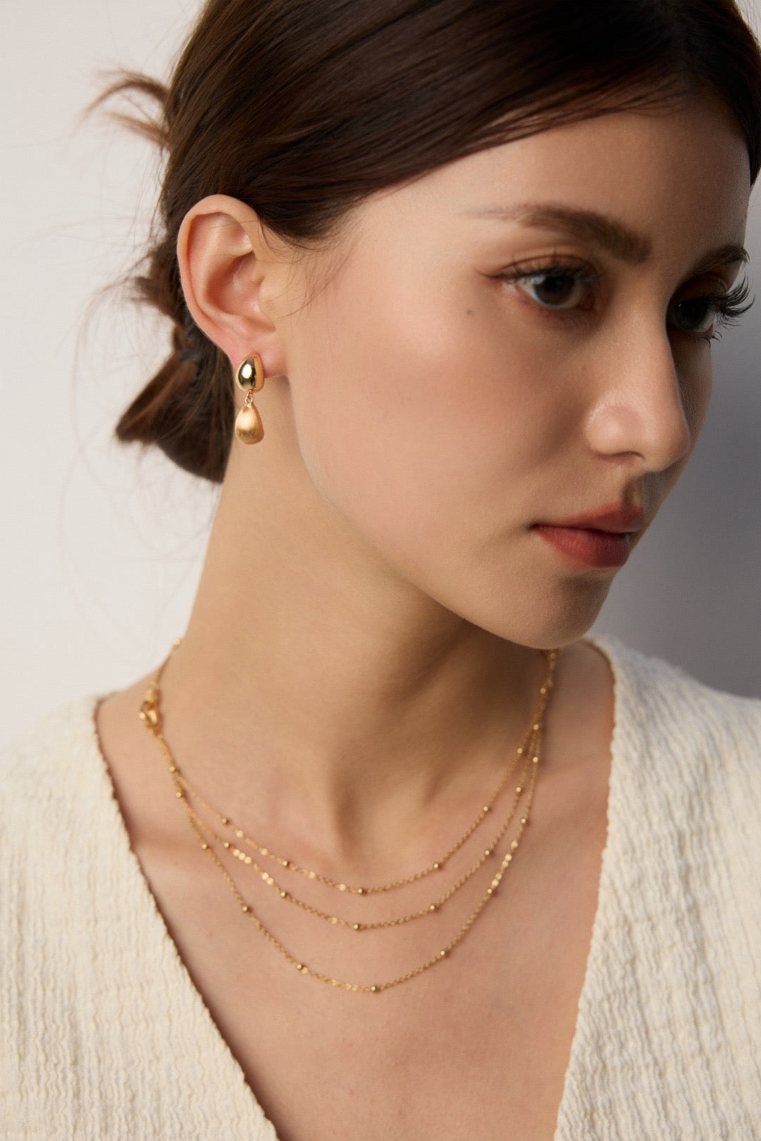 Woman wearing gold earrings and necklaces against a neutral background