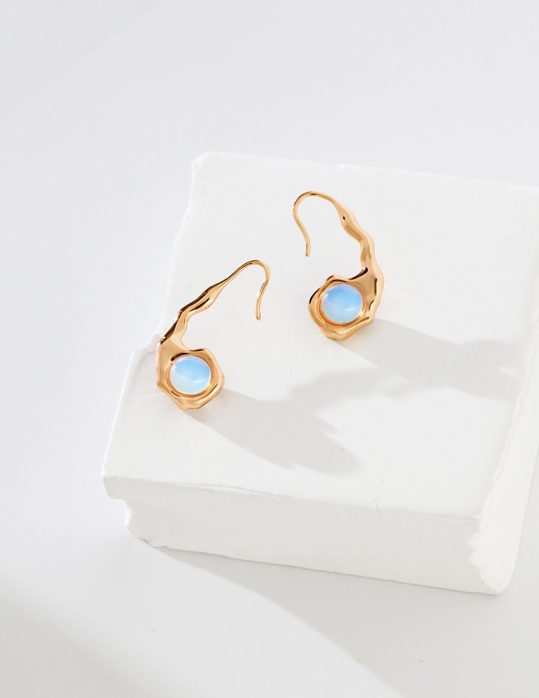 A pair of sculptural 18K gold vermeil earrings featuring a reversible design with opal on one side, displayed on a white textured block.