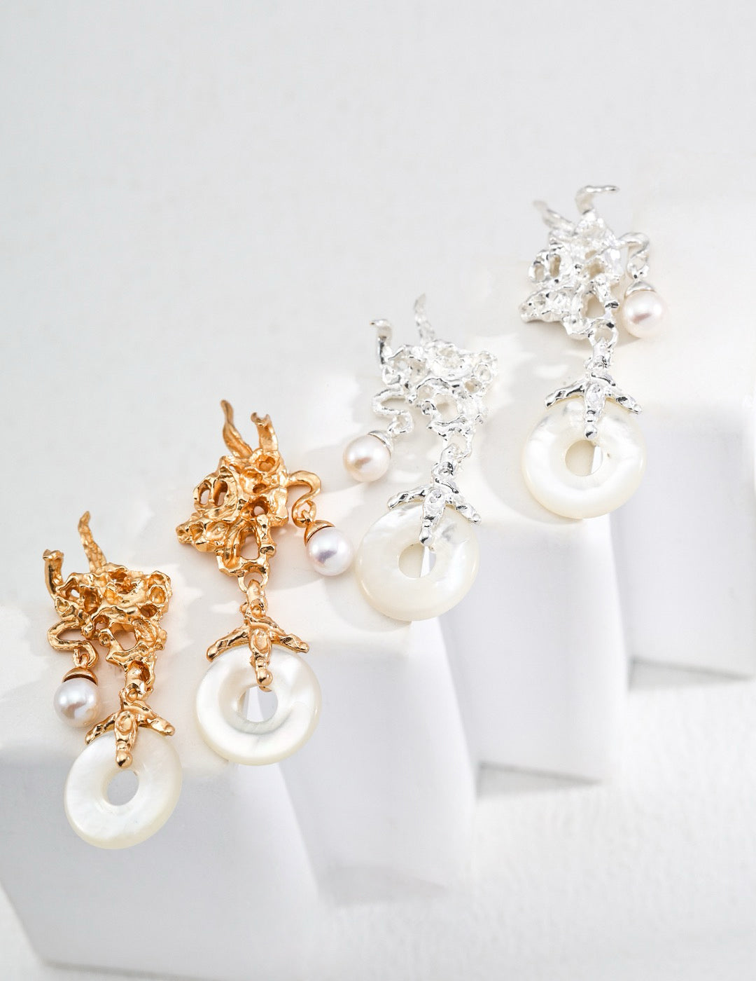 Gold and silver dangle earrings with coral-like metal design, freshwater pearls, and circular mother-of-pearl pendants, styled on a white stepped display.