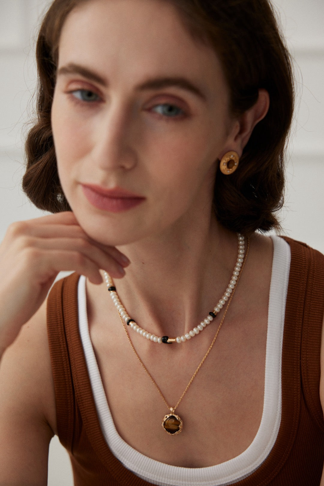 A focus shot on the model is wearing the Freshwater Pearl Black Enamel Necklace, which is layered with a Tiger's Eye Pendant Necklace, a vintage gold Tiger's Eye earring, and styled with a casual outfit.