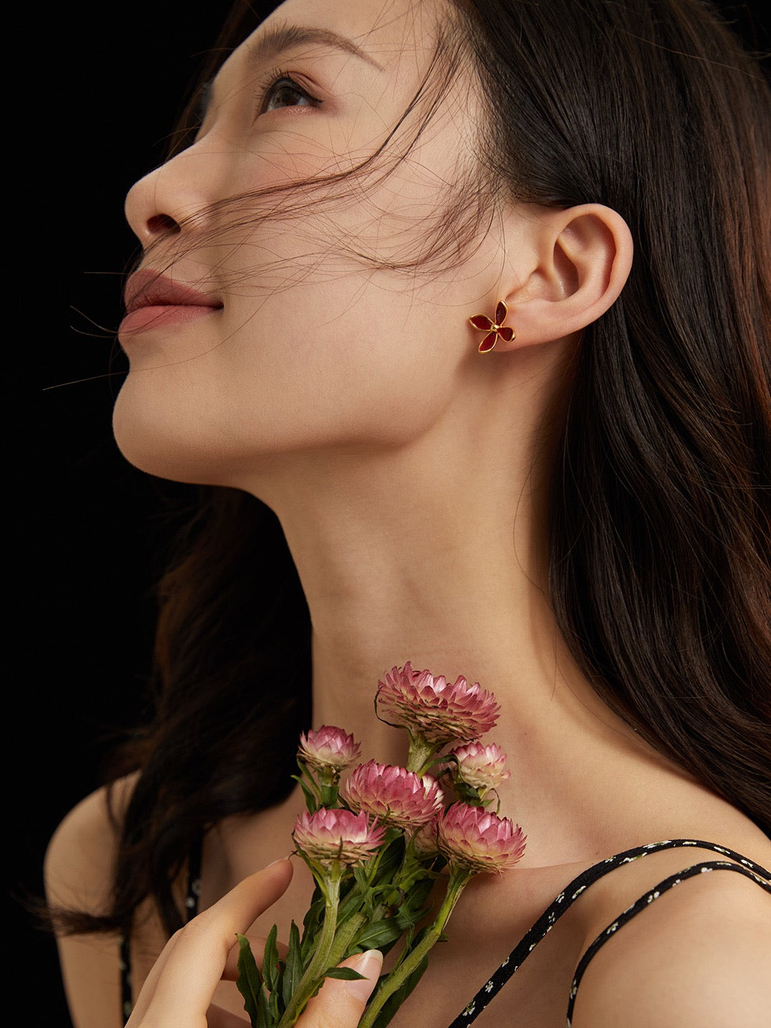 Woman wearing a red floral stud earring holding flowers against a dark background