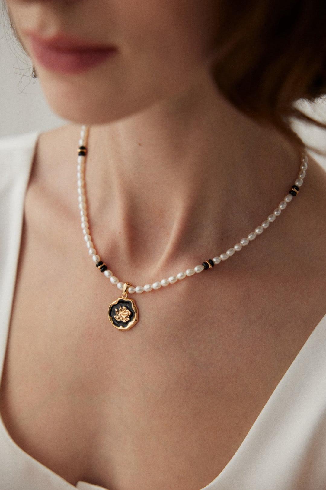 A woman wearing a white dress and the Rosanna Necklace which features black agate, pearls, and a rose-design enamel pendant.