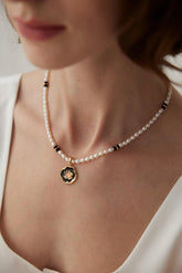 A woman wearing a white dress and the Rosanna Necklace which features black agate, pearls, and a rose-design enamel pendant.