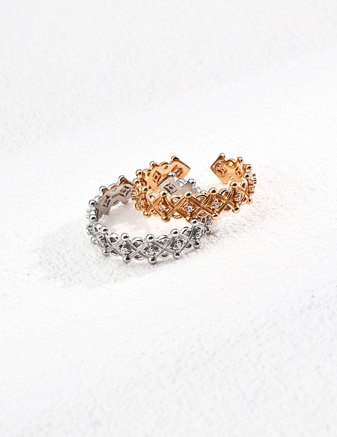 Two Royale Lattice Rings, one in gold and one in silver, featuring crisscross patterns and cubic zirconia detailing, displayed on a white textured background.