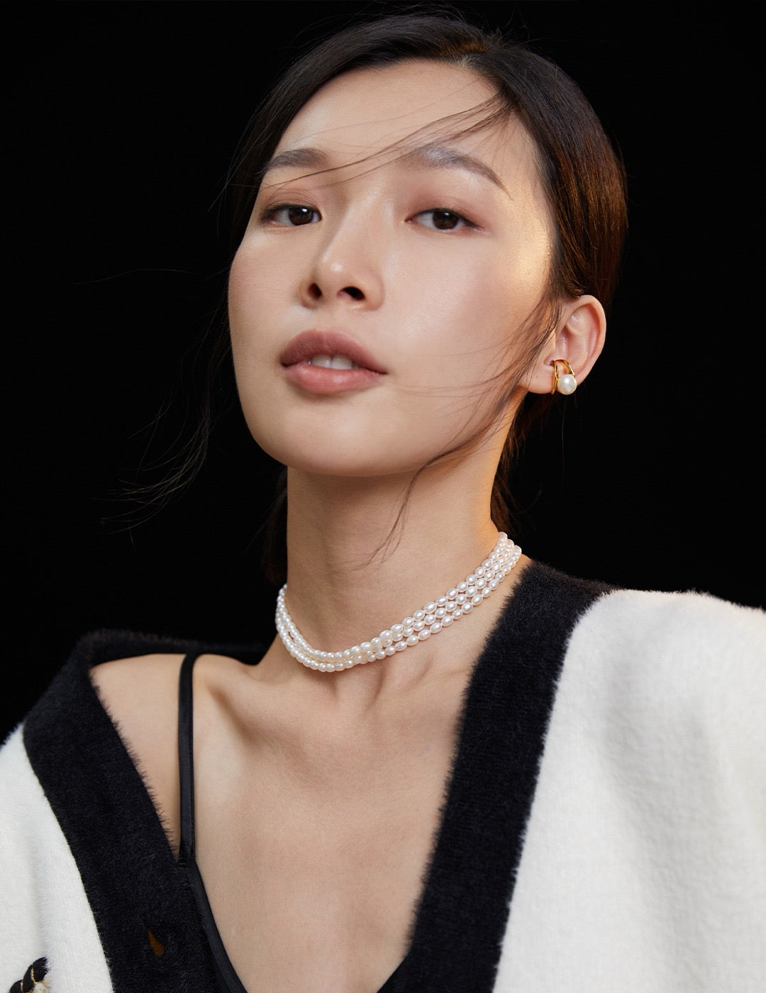 Woman wearing a black and white outfit with a triple-strand pearl choker necklace and earrings against a black background