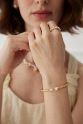 A person wearing a beige dress showcasing a gold open bangle bracelet with pearls on their wrist.