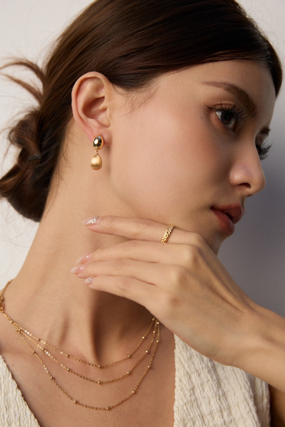 Woman wearing gold jewelry including earrings, necklace, and ring on a neutral background