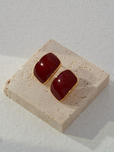 Pair of red square enamel earrings with gold accents on a beige stone surface.