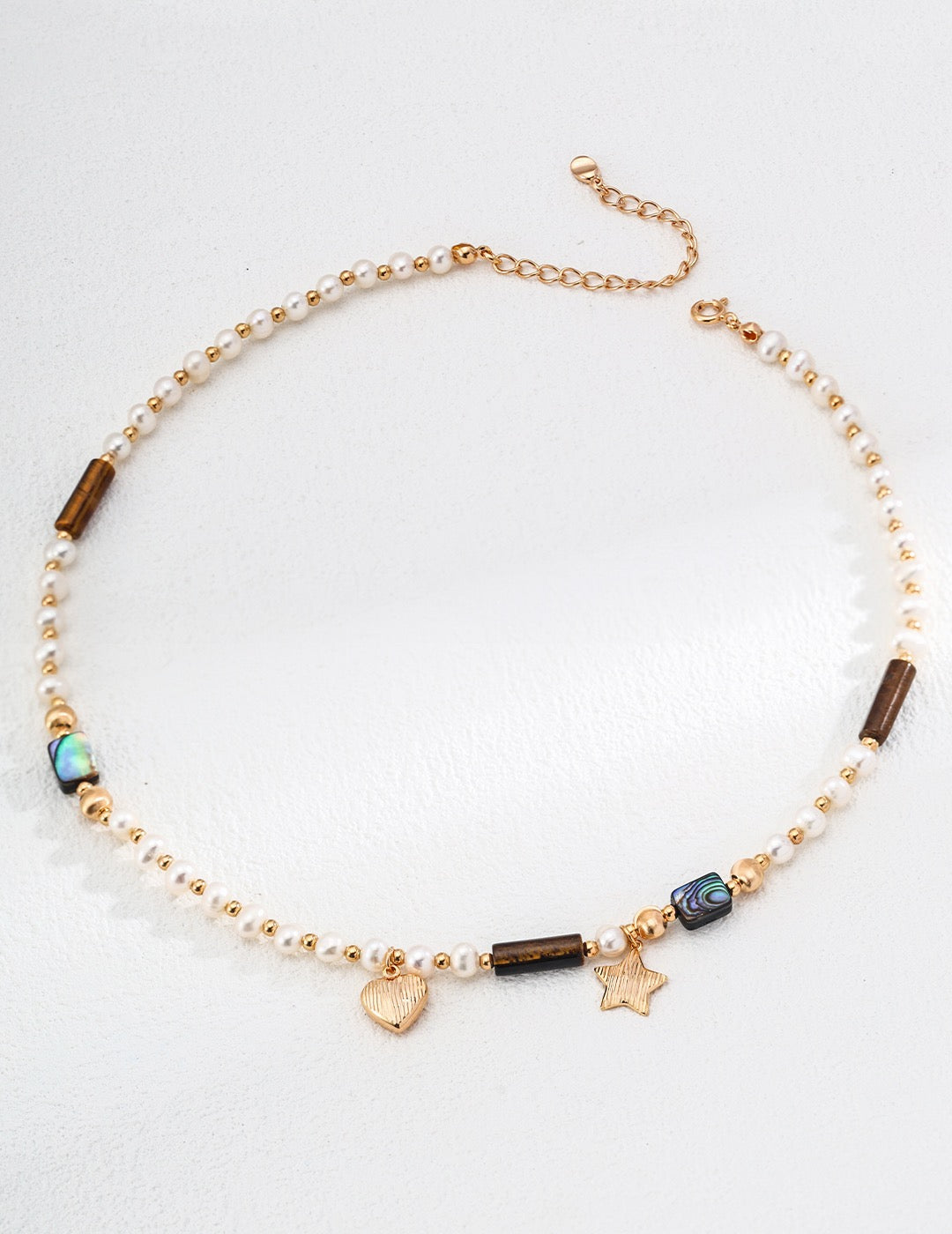 Gold-tone necklace with small freshwater pearls, tiger’s eye beads, abalone accents, and heart and star-shaped gold charms on a white background.