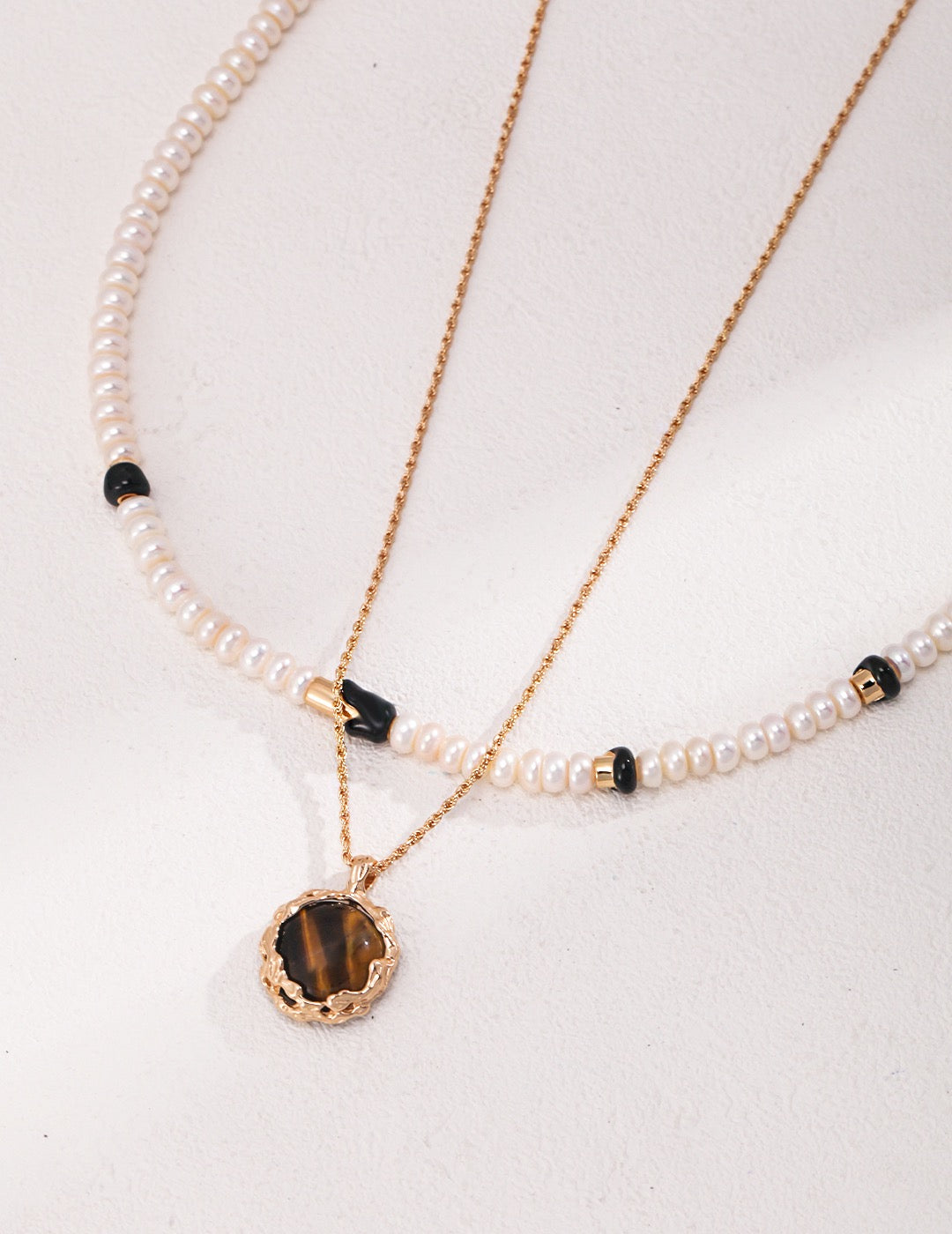 A close-up of Ceres 18K Gold Freshwater Pearl Black Enamel Necklace layered with an Tiger's Eye Pendant Necklace.