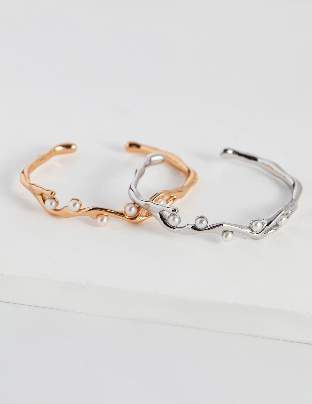 Two open cuff bracelets in gold and silver with flowing branch-like design and small white pearls, displayed on a white background.