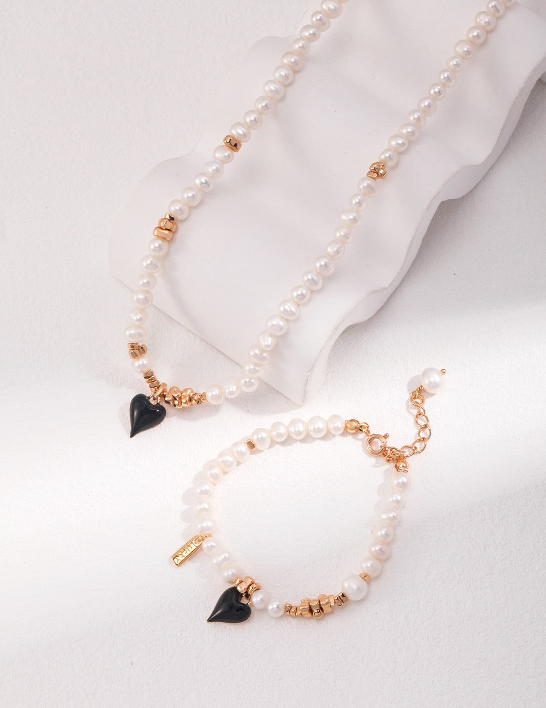 A pearl necklace and bracelet set featuring white freshwater pearls, gold accents, and black heart-shaped charms, styled on a soft white surface with gentle natural lighting.