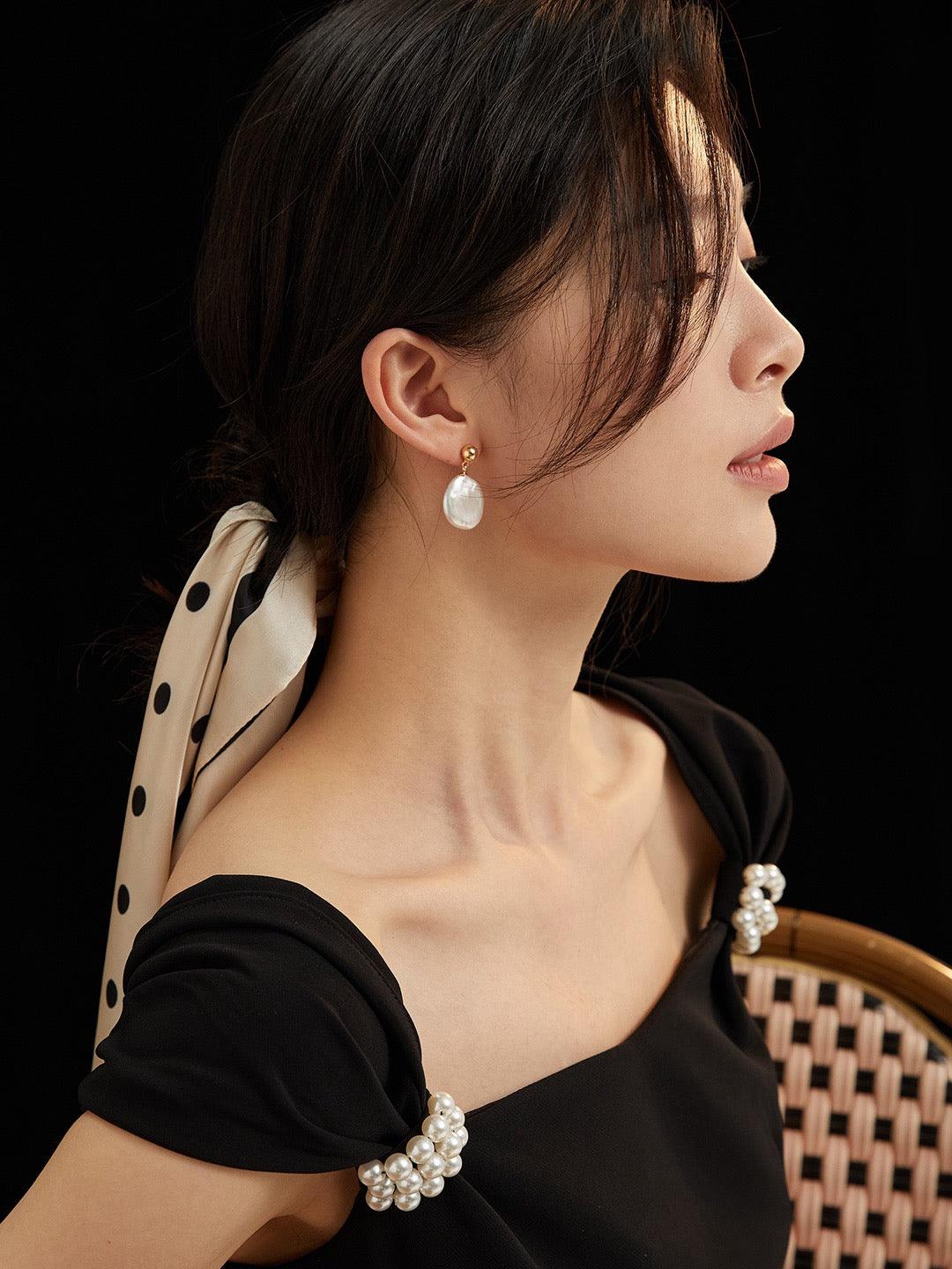 A woman wearing Aila Baroque Pearl Drop Earrings with 18K Gold Vermeil, paired with a black top and a polka dot accessory.