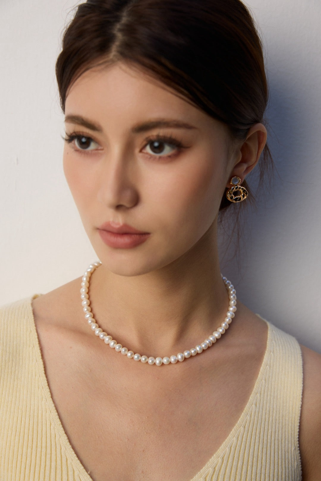 Woman wearing a 7mm pearl strand necklace and gold earrings against a plain background
