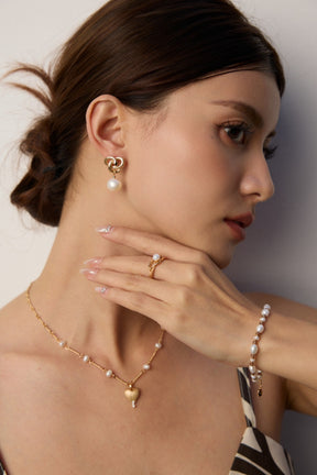 Woman wearing gold pearl jewelry, including drop earrings, necklace, ring, and bra,celet against a neutral background