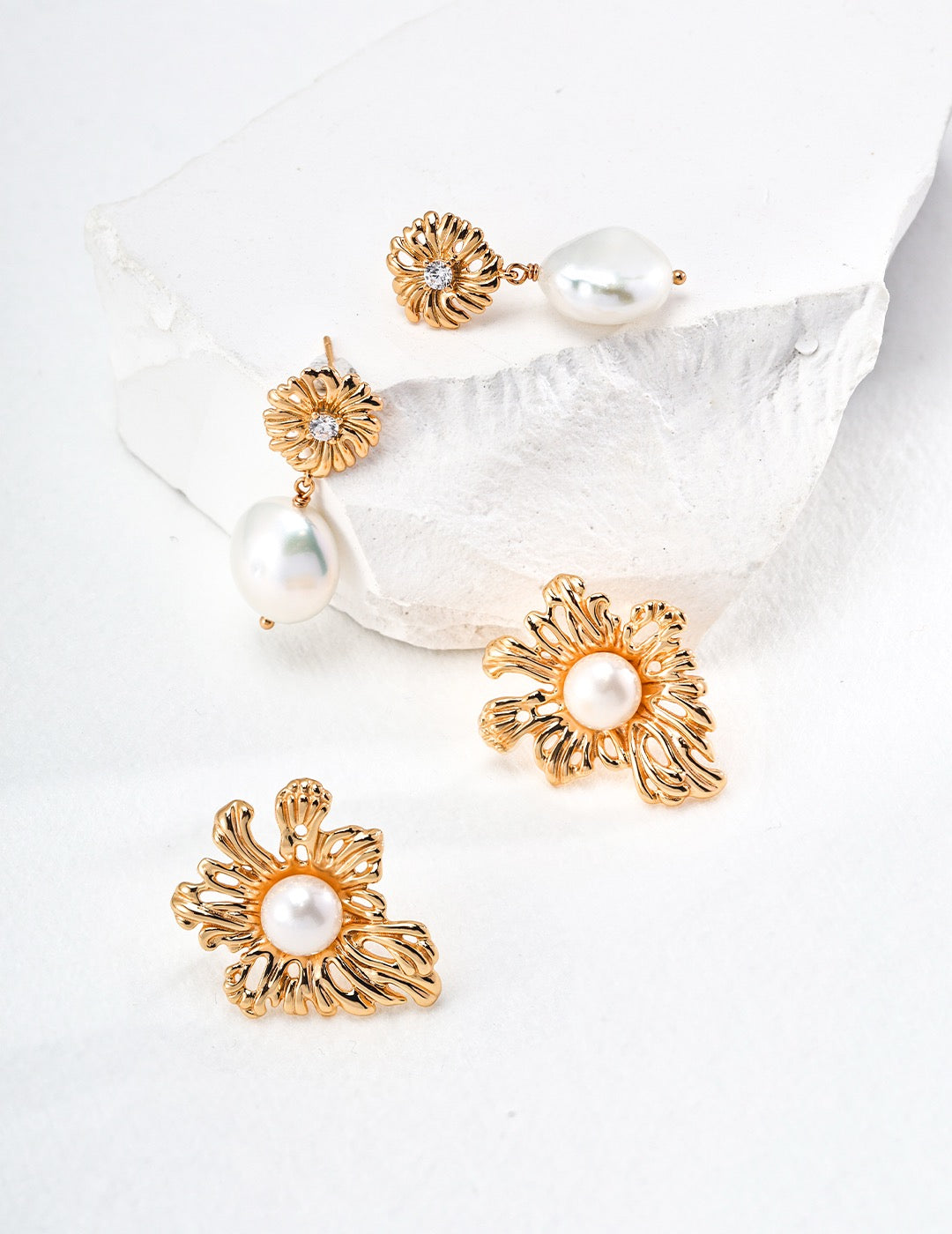 A pair of gold floral pearl earrings displayed on a white surface, with one earring slightly in front of the other, creating a dynamic sense of depth.