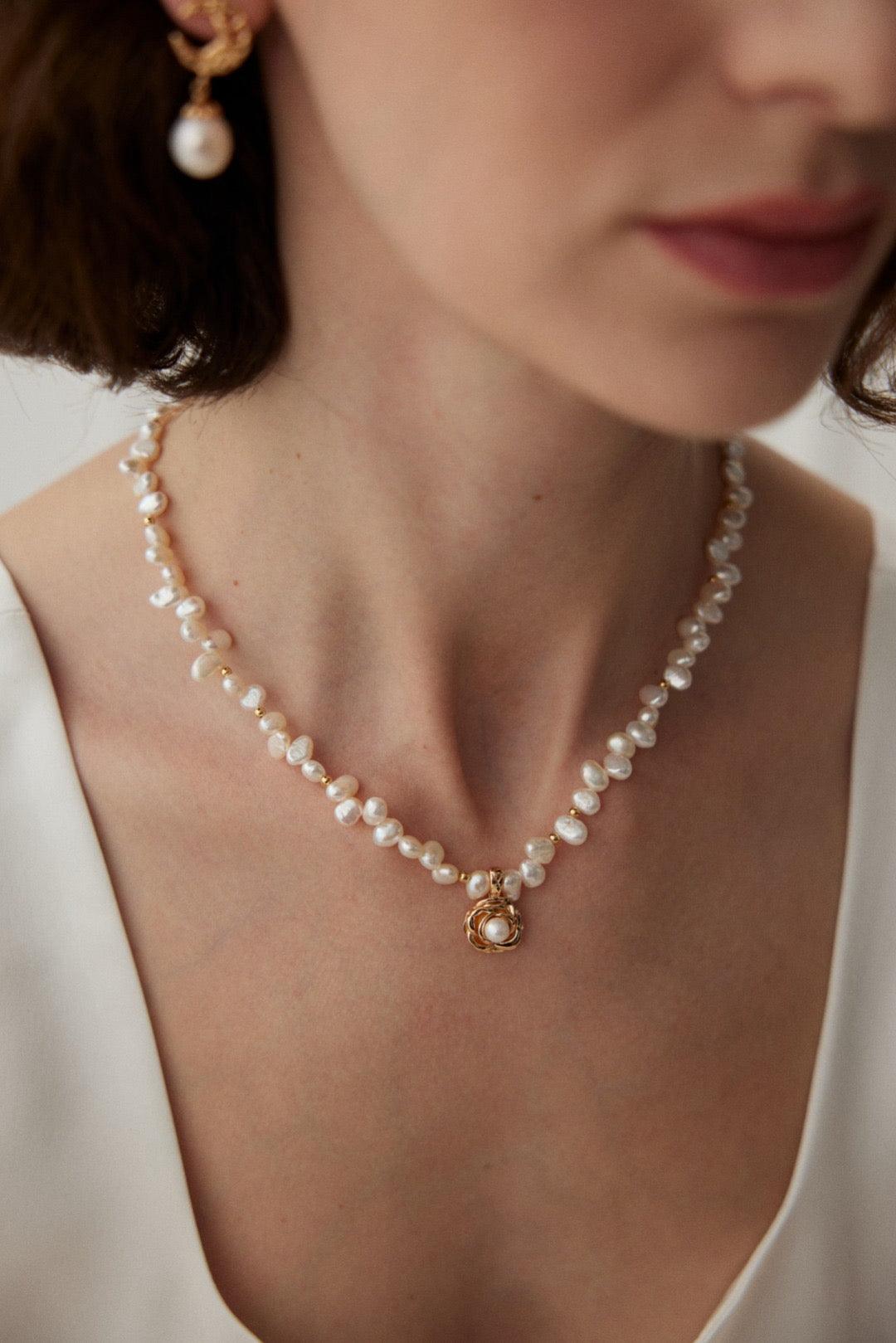 A person wearing a white garment showcasing a gold necklace with pearl beads and a camellia-shaped pendant.