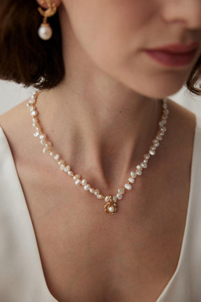 A person wearing a white garment showcasing a gold necklace with pearl beads and a camellia-shaped pendant.