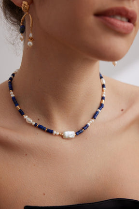 Close-up of a person wearing a necklace with blue beads and pearls, with a neutral background.