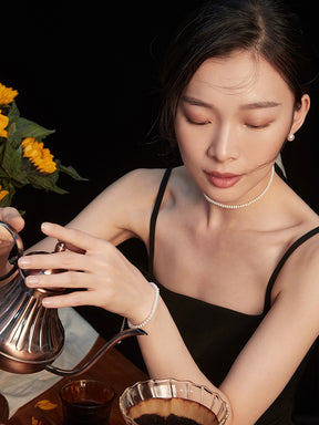 Woman is wearing pearl stud earrings and pearl choker necklace and she is pouring tea from a silver teapot with sunflowers in the background