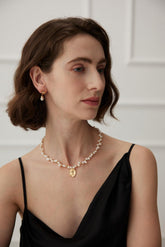 A woman wearing a sophisticated black dress and a gold-colored necklace with a round queen engraved pendant, accompanied by a string of pearls.