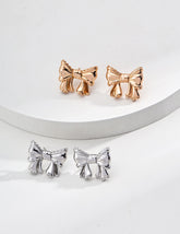 Two pairs of Coquette bow stud earrings arranged on a stepped white display