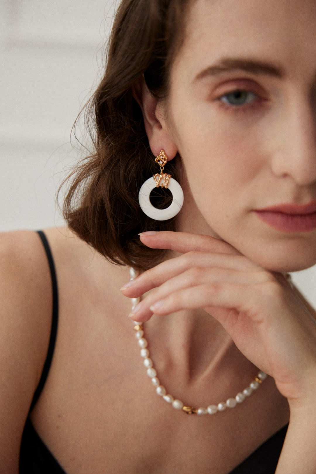 A woman wearing gold-tone filigree stud earrings with a white circular pendant drop, hypoallergenic and sustainable jewelry.