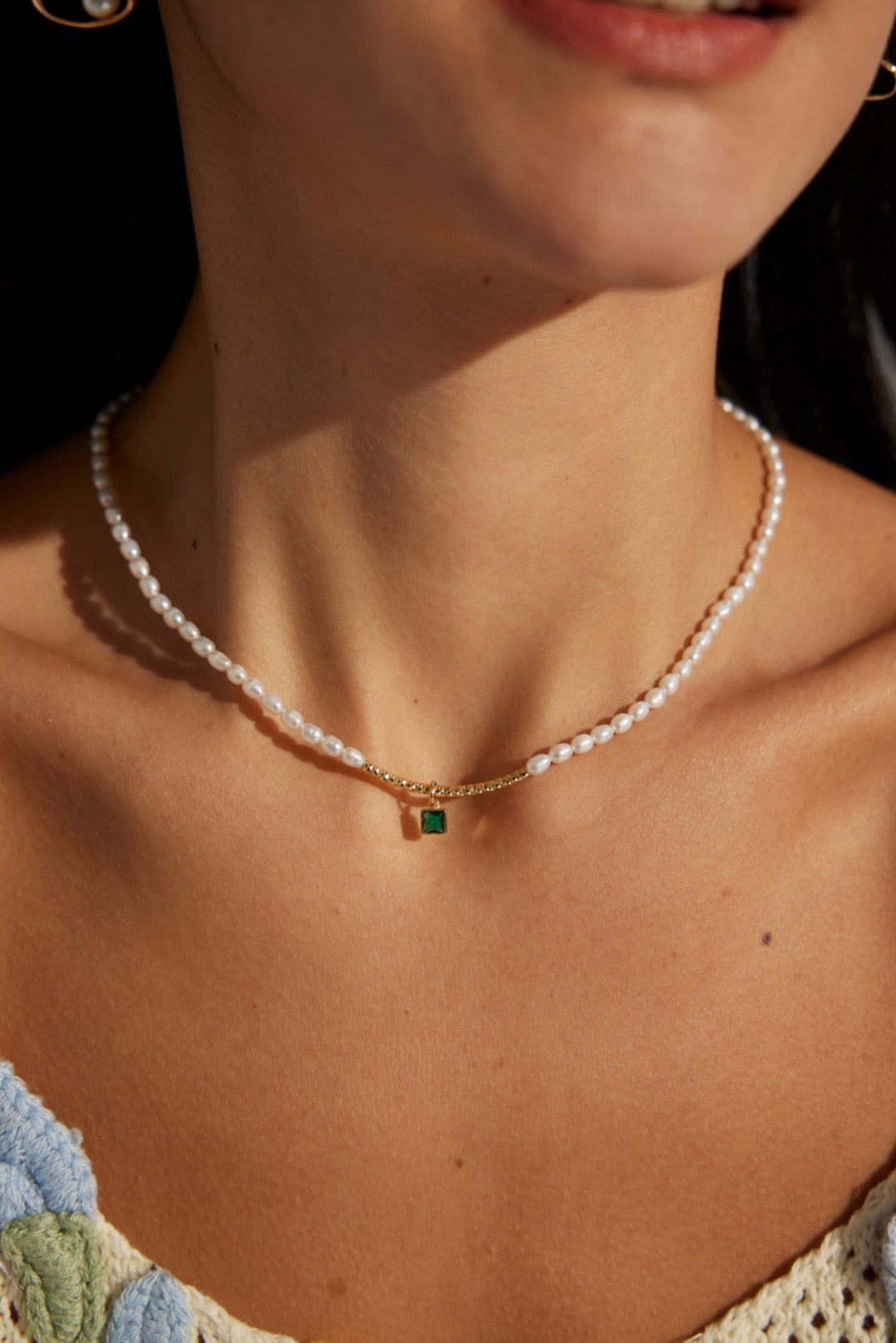 A person wearing a white, floral necklineJulia Necklace featuring white pearls and a green gemstone pendant.