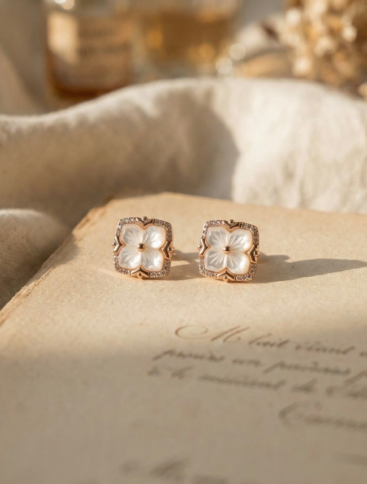 Hand-carved mother of pearl clover stud earrings with a shimmering zircon crystal halo, set in 18K gold vermeil and displayed on a vintage book.