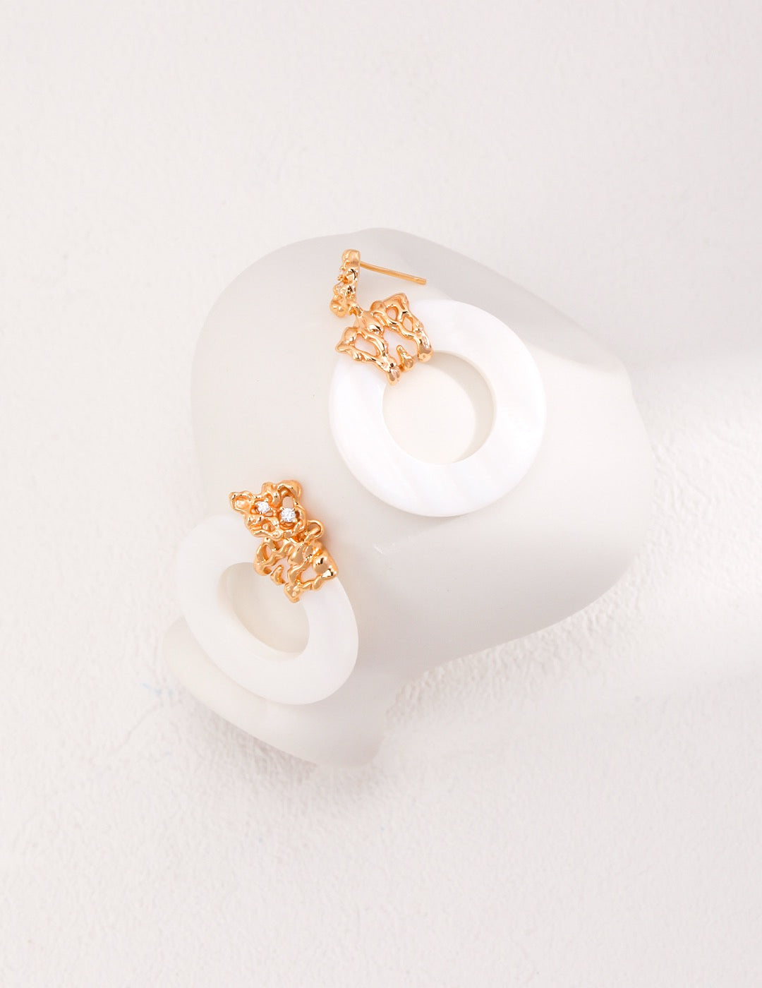 The Ethereal Harmony Hoop Earrings featuring luminous white mother-of-pearl discs suspended from intricately textured gold-toned posts, displayed on the white vassel. 