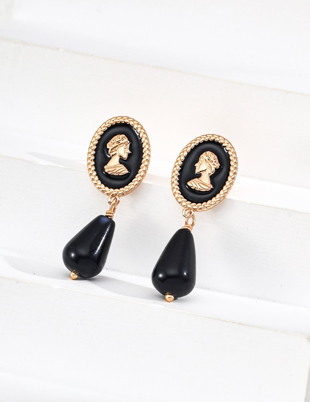 Pair of earrings with black teardrop shapes and queen cameo gold accents on a white background